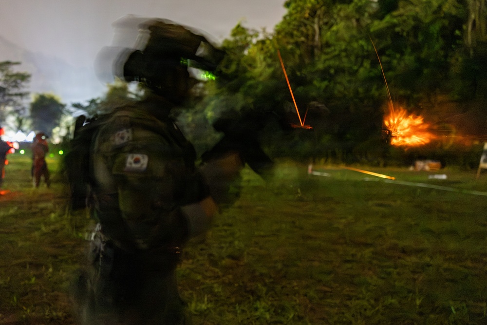 A South Korean military soldier is photographed during a nighttime live-fire training exercise. The image shows motion blur from a long exposure, capturing tracer rounds and an explosion or target detonation in the background. The soldier is equipped with full tactical gear including night vision equipment and is actively firing a weapon.