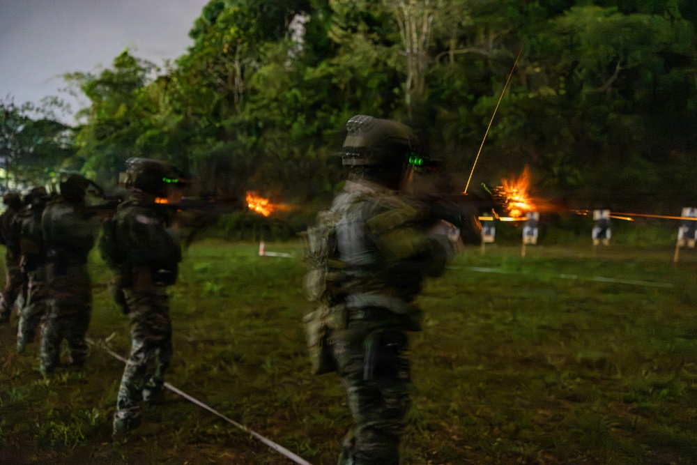 A low-light, long-exposure photograph capturing military or law enforcement personnel conducting a live-fire night shooting exercise on an outdoor range. Multiple armed figures in camouflage uniforms and helmets are visible firing weapons toward targets, with muzzle flashes and tracer rounds clearly visible. Motion blur from the long exposure gives the figures a ghostly appearance.