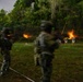 A low-light, long-exposure photograph capturing military or law enforcement personnel conducting a live-fire night shooting exercise on an outdoor range. Multiple armed figures in camouflage uniforms and helmets are visible firing weapons toward targets, with muzzle flashes and tracer rounds clearly visible. Motion blur from the long exposure gives the figures a ghostly appearance.