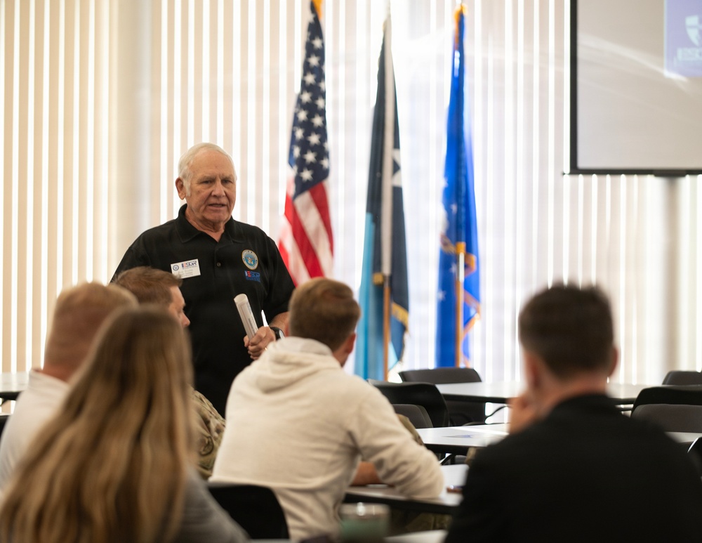 An older man in a black polo shirt is presenting or speaking to a seated audience in what appears to be a formal meeting or training room. He is holding papers and wearing name badges/lanyards, standing in front of a row of flags including the American flag and what appear to be military or organizational flags. A projection screen is visible in the upper right corner.