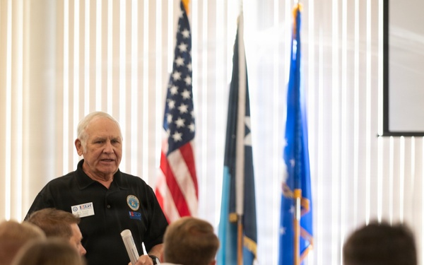 An older man in a black polo shirt is presenting or speaking to a seated audience in what appears to be a formal meeting or training room. He is holding papers and wearing name badges/lanyards, standing in front of a row of flags including the American flag and what appear to be military or organizational flags. A projection screen is visible in the upper right corner.