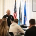 An older man in a black polo shirt is presenting or speaking to a seated audience in what appears to be a formal meeting or training room. He is holding papers and wearing name badges/lanyards, standing in front of a row of flags including the American flag and what appear to be military or organizational flags. A projection screen is visible in the upper right corner.