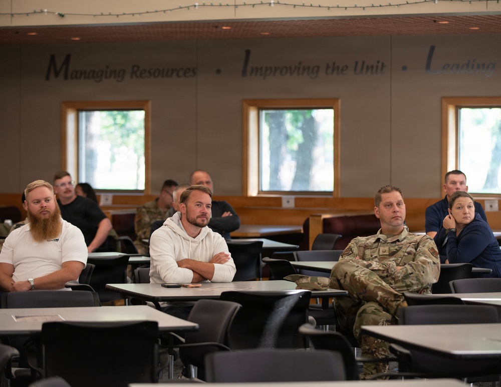 A group of individuals, including at least one person in military camouflage uniform, are seated at desks in a classroom or briefing room setting. They appear to be attending a presentation or training session, with most participants facing forward attentively. The room displays motivational text on the wall reading 'Managing Resources • Improving the Unit • Leading...'