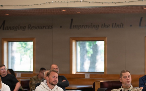 A group of individuals, including at least one person in military camouflage uniform, are seated at desks in a classroom or briefing room setting. They appear to be attending a presentation or training session, with most participants facing forward attentively. The room displays motivational text on the wall reading 'Managing Resources • Improving the Unit • Leading...'