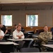 A group of individuals, including at least one person in military camouflage uniform, are seated at desks in a classroom or briefing room setting. They appear to be attending a presentation or training session, with most participants facing forward attentively. The room displays motivational text on the wall reading 'Managing Resources • Improving the Unit • Leading...'