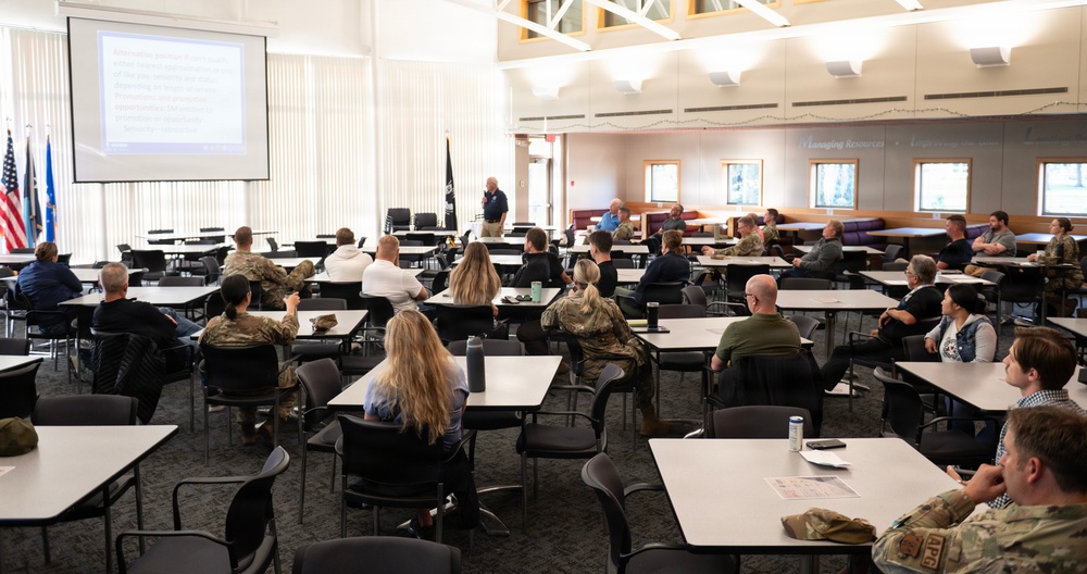 A briefing or training session is taking place in a large conference or dining facility, with a presenter standing at the front using a projected presentation on a screen. The audience consists of a mix of military personnel in camouflage uniforms and civilians seated at individual tables. The setting appears to be on a military installation based on the flags and attire of attendees.