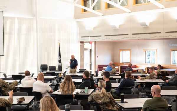 A briefing or training session is taking place in a large conference or dining facility, with a presenter standing at the front using a projected presentation on a screen. The audience consists of a mix of military personnel in camouflage uniforms and civilians seated at individual tables. The setting appears to be on a military installation based on the flags and attire of attendees.