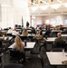A briefing or training session is taking place in a large conference or dining facility, with a presenter standing at the front using a projected presentation on a screen. The audience consists of a mix of military personnel in camouflage uniforms and civilians seated at individual tables. The setting appears to be on a military installation based on the flags and attire of attendees.