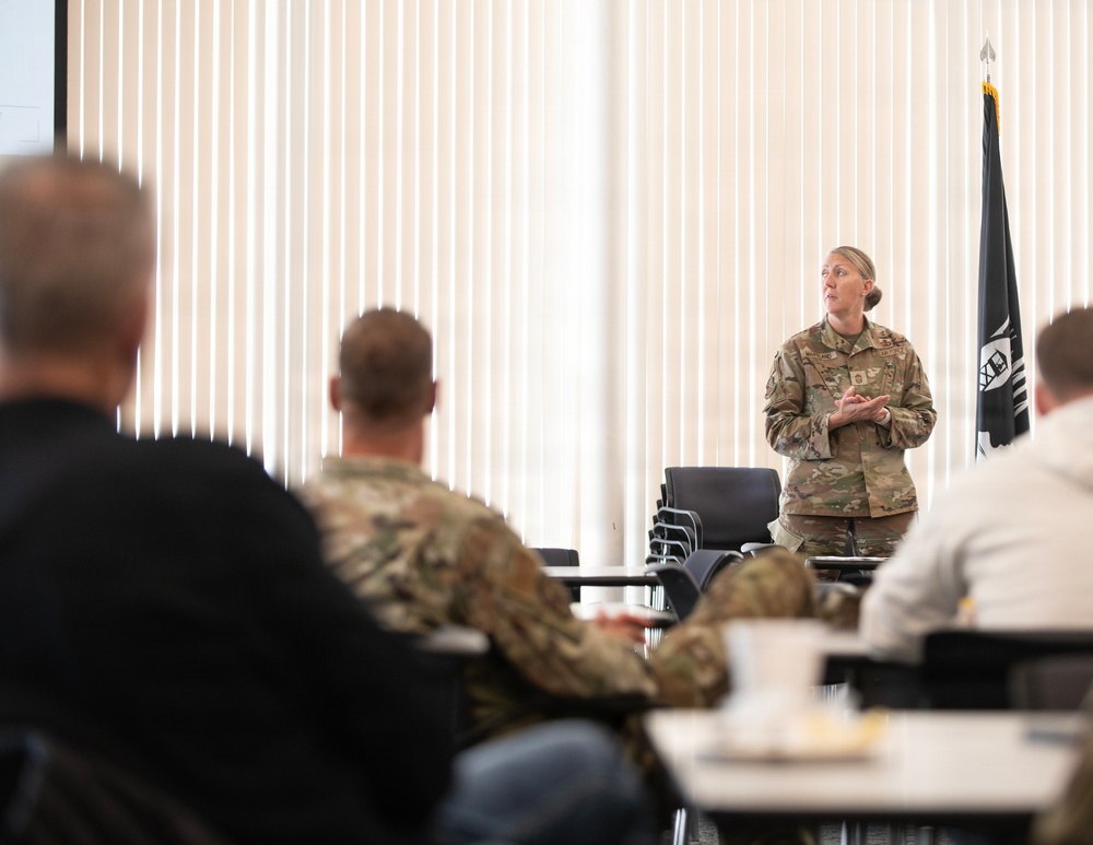 A female military officer in OCP (Operational Camouflage Pattern) uniform is addressing a group of seated personnel in what appears to be a briefing or training room. The audience includes both uniformed military members and at least one civilian. A POW/MIA flag is visible in the background to the right of the speaker.