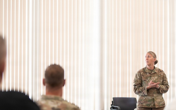 A female military officer in OCP (Operational Camouflage Pattern) uniform is addressing a group of seated personnel in what appears to be a briefing or training room. The audience includes both uniformed military members and at least one civilian. A POW/MIA flag is visible in the background to the right of the speaker.
