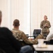 A female military officer in OCP (Operational Camouflage Pattern) uniform is addressing a group of seated personnel in what appears to be a briefing or training room. The audience includes both uniformed military members and at least one civilian. A POW/MIA flag is visible in the background to the right of the speaker.