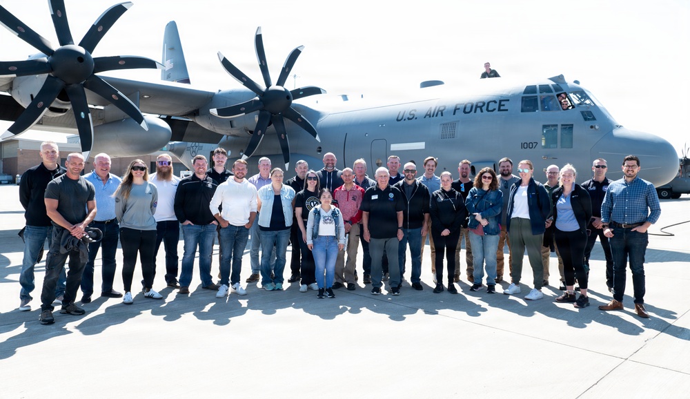 A large group of approximately 30 civilians poses for a group photo in front of a U.S. Air Force C-130J Super Hercules turboprop military transport aircraft on a tarmac. The aircraft bears the tail number 1007 and the 'U.S. AIR FORCE' marking prominently on the fuselage. The setting appears to be an official base visit or community outreach event.