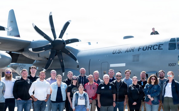 A large group of approximately 30 civilians poses for a group photo in front of a U.S. Air Force C-130J Super Hercules turboprop military transport aircraft on a tarmac. The aircraft bears the tail number 1007 and the 'U.S. AIR FORCE' marking prominently on the fuselage. The setting appears to be an official base visit or community outreach event.