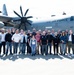 A large group of approximately 30 civilians poses for a group photo in front of a U.S. Air Force C-130J Super Hercules turboprop military transport aircraft on a tarmac. The aircraft bears the tail number 1007 and the 'U.S. AIR FORCE' marking prominently on the fuselage. The setting appears to be an official base visit or community outreach event.