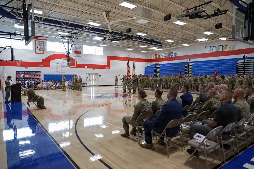 A military ceremony is taking place inside a school gymnasium identified by 'Rockets' branding with red, white, and blue colors. Numerous uniformed military personnel are assembled in formation while seated dignitaries and officers observe a speaker at a podium. The event appears to be a formal military function such as a change of command, activation, or deployment ceremony.