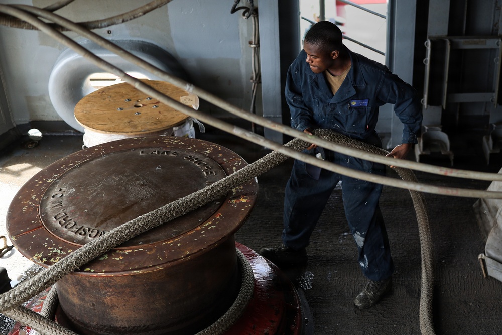 A sailor or maritime worker in dark blue coveralls is handling thick mooring ropes on what appears to be the deck of a large vessel. He is actively pulling or managing heavy braided lines around a large capstan or bollard. The setting is an industrial ship deck with various maritime equipment visible.