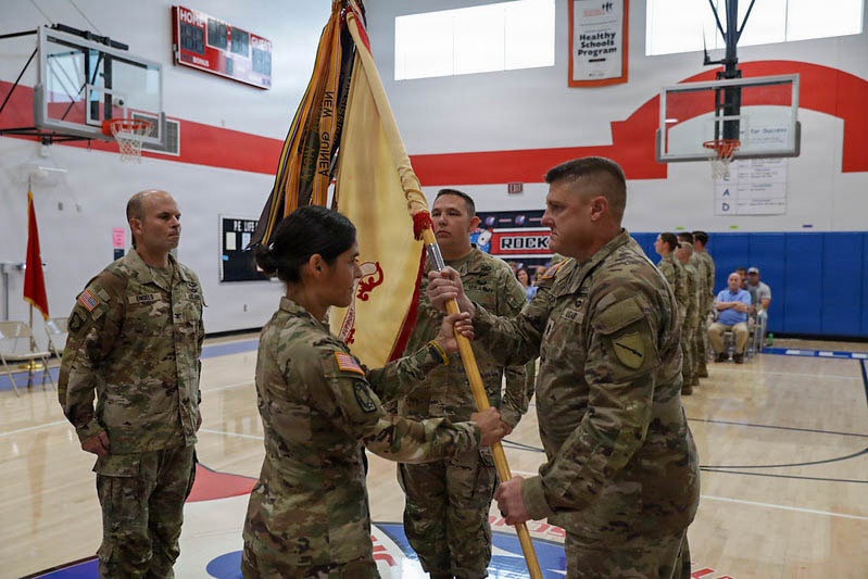 A U.S. Army change of command ceremony is taking place inside a school gymnasium. A female soldier is passing a unit guidon (flag) to a male soldier, which is the central symbolic act of a change of command. Additional soldiers are standing in formation along the sides of the gym, with civilian observers seated in the background.