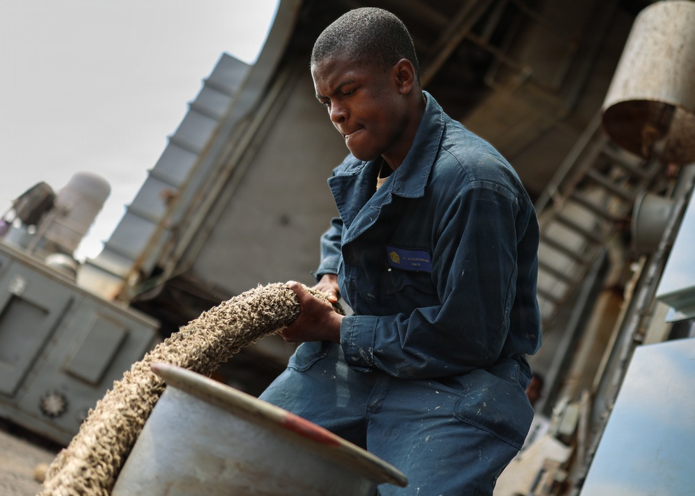 A young man wearing blue coveralls with a name tag is handling a thick rope or hawser, coiling or managing it into a large metal container. The setting appears to be an industrial or maritime environment, likely the deck of a ship or dock area. The image is taken from a low angle, emphasizing the subject's focused expression and physical effort.