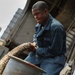 A young man wearing blue coveralls with a name tag is handling a thick rope or hawser, coiling or managing it into a large metal container. The setting appears to be an industrial or maritime environment, likely the deck of a ship or dock area. The image is taken from a low angle, emphasizing the subject's focused expression and physical effort.