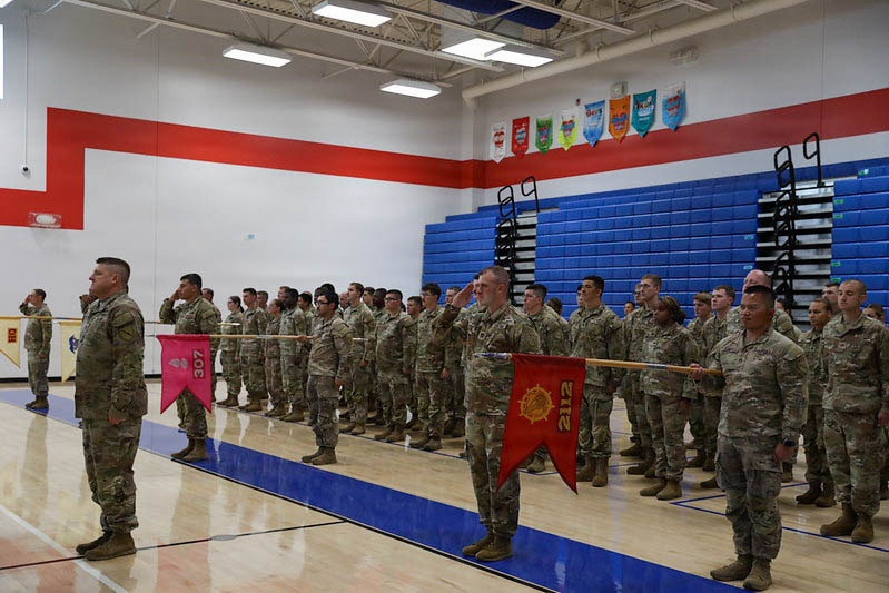 A military formation ceremony is taking place inside a gymnasium, with soldiers in Army Combat Uniforms (ACUs) standing at attention and saluting. Two unit guidons are visible, bearing the numbers '307' and '212', indicating distinct military units are represented. The setting appears to be a school or recreational facility gymnasium with blue bleachers and red/white/blue wall markings.
