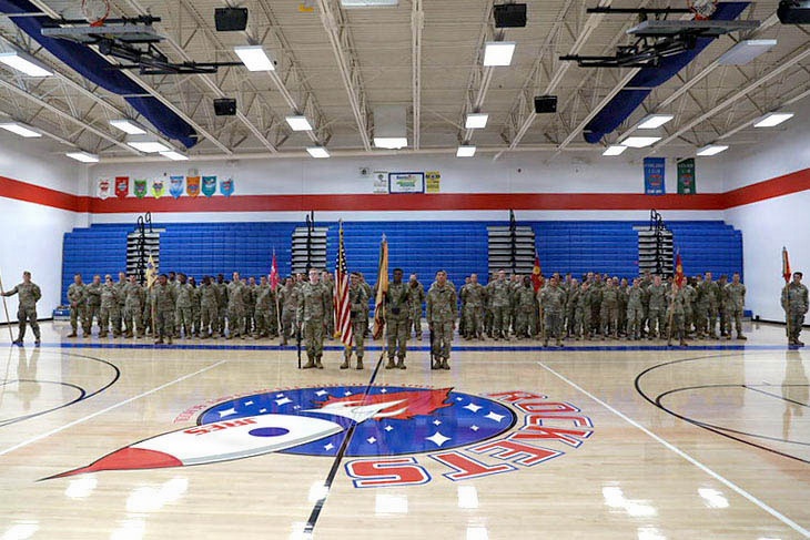 A large group of U.S. Army soldiers in camouflage uniforms are assembled in formation inside a school gymnasium. The formation includes color guard personnel holding the American flag and what appear to be unit guidons at the center. The gym features red, white, and blue décor with a 'Rockets' mascot logo on the court floor.