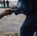 A person in blue work coveralls is gripping and pulling a large, weathered mooring rope on what appears to be a ship's deck or dock. The coveralls show paint stains and wear, suggesting active maritime or naval work. Several other individuals are visible in the blurred background, indicating a group operation such as a ship docking or undocking procedure.