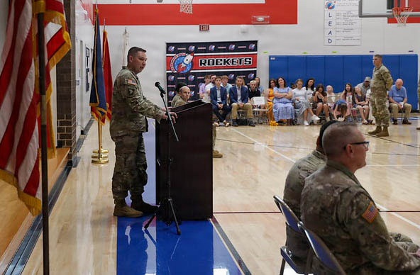 A military ceremony is taking place in a school gymnasium identified as belonging to the 'Rockets' team. A soldier in Army Combat Uniform (ACU) is speaking at a podium while other uniformed personnel and civilian attendees are seated as an audience. The setting includes American and military flags, suggesting a formal military event such as a promotion, award, or deployment ceremony.