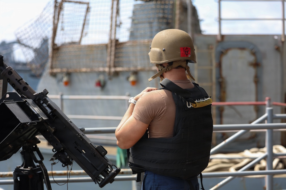 A military or naval personnel member is standing on the deck of a vessel wearing a tan combat helmet and a black body armor vest with 'US' markings visible. The individual is facing away from the camera with arms crossed, positioned next to a mounted crew-served weapon, likely an M2 .50 caliber machine gun. A second gray naval vessel is visible in the background.