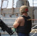 A military or naval personnel member is standing on the deck of a vessel wearing a tan combat helmet and a black body armor vest with 'US' markings visible. The individual is facing away from the camera with arms crossed, positioned next to a mounted crew-served weapon, likely an M2 .50 caliber machine gun. A second gray naval vessel is visible in the background.