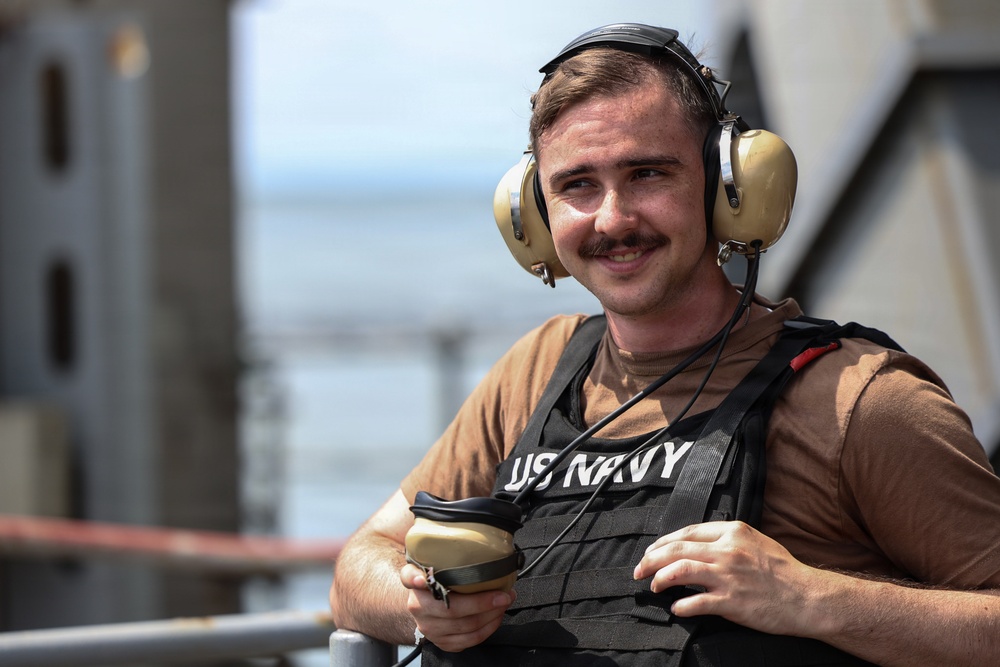 A young male US Navy personnel is photographed outdoors, likely on the deck of a naval vessel. He is smiling at the camera while wearing standard naval working gear including hearing protection and a tactical vest. The background shows blurred structural elements consistent with a ship's superstructure and open water.