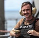 A young male US Navy personnel is photographed outdoors, likely on the deck of a naval vessel. He is smiling at the camera while wearing standard naval working gear including hearing protection and a tactical vest. The background shows blurred structural elements consistent with a ship's superstructure and open water.