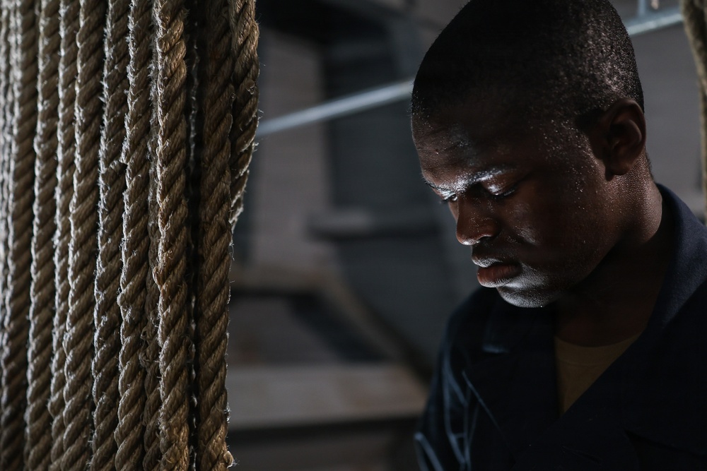 A close-up portrait of a man with visible perspiration on his face, positioned next to thick natural fiber ropes. The dramatic lighting and sweat suggest physical exertion in what appears to be an industrial or athletic setting. The composition emphasizes the man's focused, downward gaze and the textural contrast between his skin and the rough ropes.