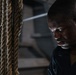 A close-up portrait of a man with visible perspiration on his face, positioned next to thick natural fiber ropes. The dramatic lighting and sweat suggest physical exertion in what appears to be an industrial or athletic setting. The composition emphasizes the man's focused, downward gaze and the textural contrast between his skin and the rough ropes.