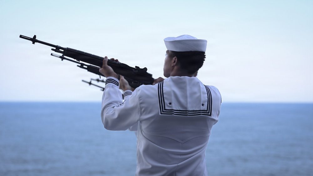 A naval sailor in dress white uniform is shown from behind, raising a rifle skyward over an open ocean backdrop. The setting and posture suggest a ceremonial rifle salute, consistent with military funeral honors or a commemorative ceremony at sea. The image is well-composed with a calm ocean horizon providing a solemn backdrop.