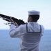 A naval sailor in dress white uniform is shown from behind, raising a rifle skyward over an open ocean backdrop. The setting and posture suggest a ceremonial rifle salute, consistent with military funeral honors or a commemorative ceremony at sea. The image is well-composed with a calm ocean horizon providing a solemn backdrop.
