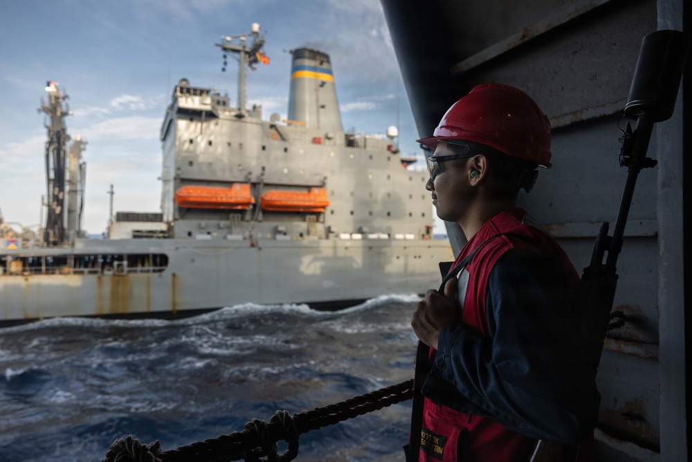 A naval or maritime worker wearing a red hard hat, safety glasses, ear protection, and a red safety vest stands on the deck of a vessel, observing a large gray replenishment or auxiliary ship nearby. The two vessels appear to be conducting or preparing for an underway replenishment (UNREP) operation at sea. The image captures the operational environment with rough water visible between the two ships.