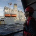 A naval or maritime worker wearing a red hard hat, safety glasses, ear protection, and a red safety vest stands on the deck of a vessel, observing a large gray replenishment or auxiliary ship nearby. The two vessels appear to be conducting or preparing for an underway replenishment (UNREP) operation at sea. The image captures the operational environment with rough water visible between the two ships.
