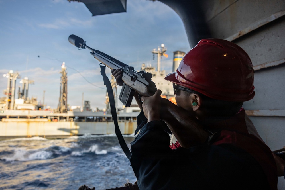 A person wearing a red hard hat, safety glasses, and ear protection is aiming a rifle with a suppressor/muzzle device from the deck of a vessel. The setting appears to be a maritime environment with industrial port infrastructure and another large ship visible in the background. The individual appears to be in a security or military role aboard the vessel.