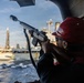 A person wearing a red hard hat, safety glasses, and ear protection is aiming a rifle with a suppressor/muzzle device from the deck of a vessel. The setting appears to be a maritime environment with industrial port infrastructure and another large ship visible in the background. The individual appears to be in a security or military role aboard the vessel.
