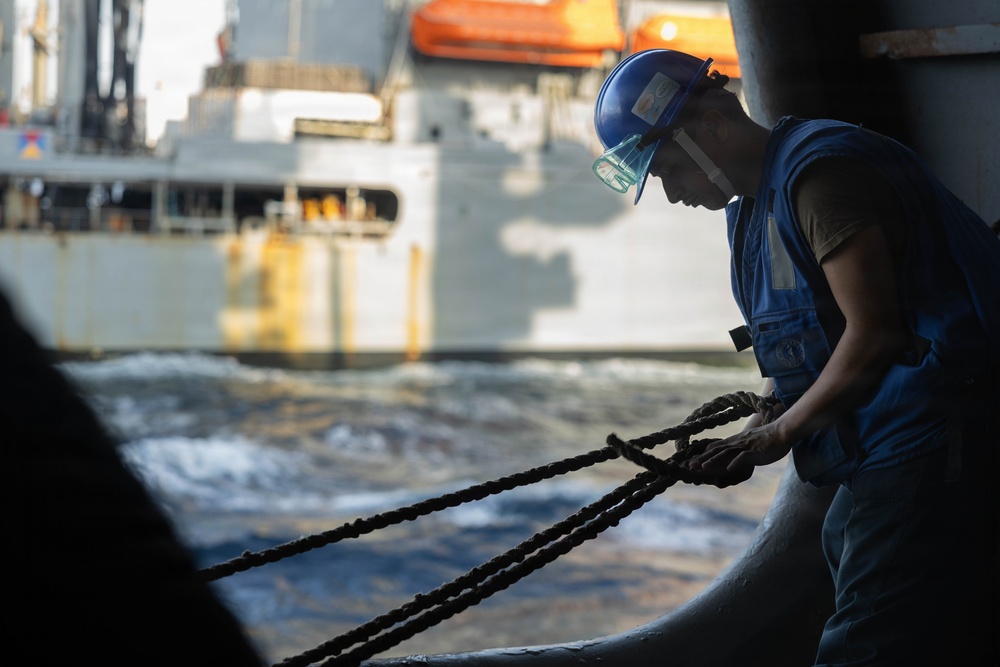 A maritime worker wearing a blue hard hat, safety goggles, and a blue vest is handling thick mooring ropes on the deck of a vessel. Another large ship is visible in the background across churning water, suggesting an underway replenishment or docking operation. The image is taken from a low angle with dramatic side lighting emphasizing the physical effort of the task.