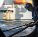 A maritime worker wearing a blue hard hat, safety goggles, and a blue vest is handling thick mooring ropes on the deck of a vessel. Another large ship is visible in the background across churning water, suggesting an underway replenishment or docking operation. The image is taken from a low angle with dramatic side lighting emphasizing the physical effort of the task.