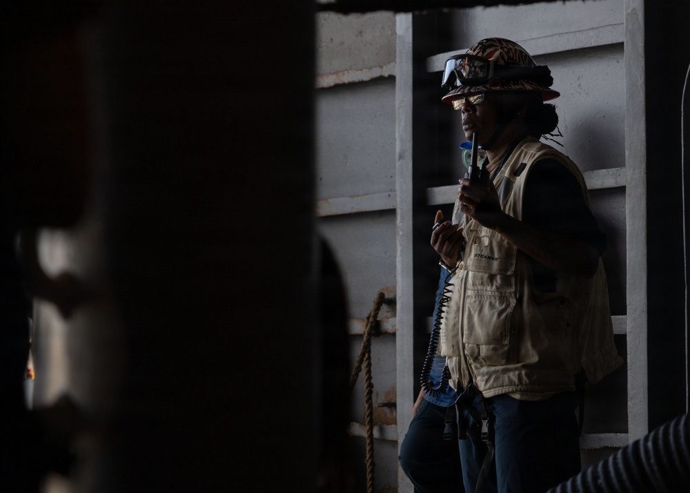 A worker wearing a hard hat with attached goggles and a tan/khaki safety vest is holding a handheld radio (walkie-talkie) in what appears to be an industrial or construction environment. The image is taken from a partially obscured angle with dark foreground elements framing the subject. The setting appears to be inside or adjacent to a large metal structure.