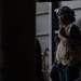 A worker wearing a hard hat with attached goggles and a tan/khaki safety vest is holding a handheld radio (walkie-talkie) in what appears to be an industrial or construction environment. The image is taken from a partially obscured angle with dark foreground elements framing the subject. The setting appears to be inside or adjacent to a large metal structure.