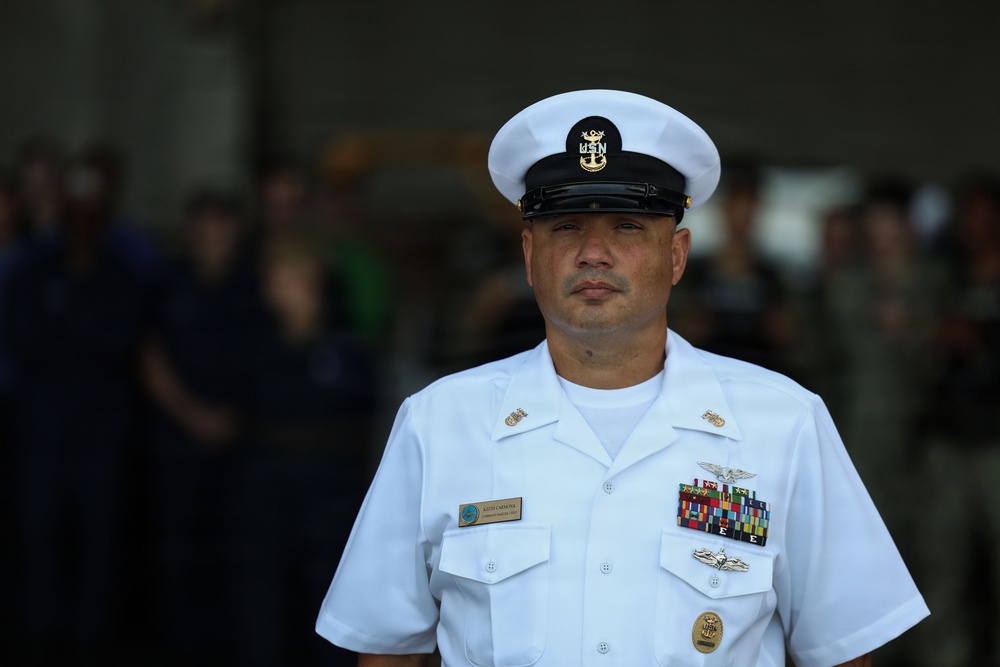 A U.S. Navy senior enlisted sailor stands at attention in dress white uniform during what appears to be a formal military ceremony. The subject displays multiple service ribbons, warfare qualification badges, and chief petty officer insignia. The background shows a blurred crowd of attendees in an indoor or covered outdoor venue.