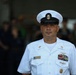A U.S. Navy senior enlisted sailor stands at attention in dress white uniform during what appears to be a formal military ceremony. The subject displays multiple service ribbons, warfare qualification badges, and chief petty officer insignia. The background shows a blurred crowd of attendees in an indoor or covered outdoor venue.