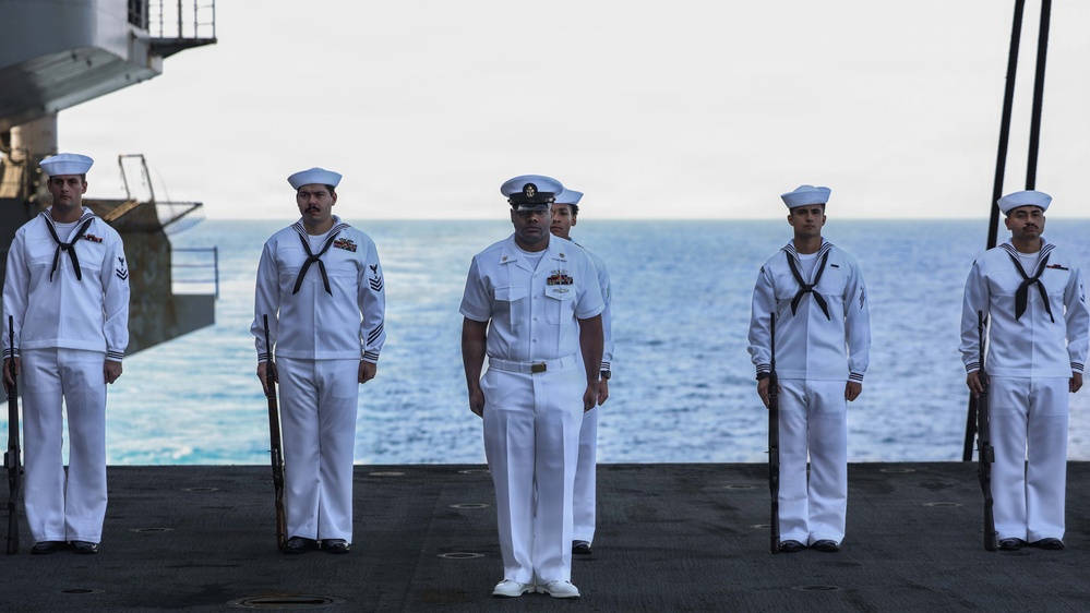 A group of U.S. Navy personnel in white dress uniforms are standing at attention on the flight deck of a naval vessel at sea. A central figure wearing a distinct officer/chief petty officer uniform with a peaked cap stands in front, while enlisted sailors holding rifles flank him on both sides. The formation appears to be part of a formal ceremony, likely a burial at sea or honors ceremony.