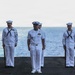 A group of U.S. Navy personnel in white dress uniforms are standing at attention on the flight deck of a naval vessel at sea. A central figure wearing a distinct officer/chief petty officer uniform with a peaked cap stands in front, while enlisted sailors holding rifles flank him on both sides. The formation appears to be part of a formal ceremony, likely a burial at sea or honors ceremony.