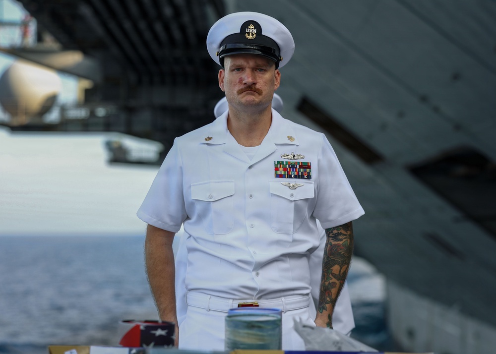 A U.S. Navy senior enlisted sailor in dress white uniform stands at attention on what appears to be the deck of a naval vessel. He is wearing a combination cover (white cap) with a USN insignia, multiple service ribbons, warfare qualification badges, and has visible tattoos on his right arm. The setting suggests a formal ceremony or official event aboard a ship.