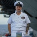 A U.S. Navy senior enlisted sailor in dress white uniform stands at attention on what appears to be the deck of a naval vessel. He is wearing a combination cover (white cap) with a USN insignia, multiple service ribbons, warfare qualification badges, and has visible tattoos on his right arm. The setting suggests a formal ceremony or official event aboard a ship.