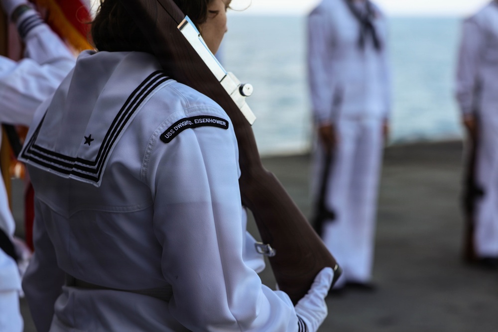 A U.S. Navy sailor in dress white uniform is shown from behind, wearing a shoulder patch reading 'USS DWIGHT D. EISENHOWER'. The sailor appears to be participating in a formal military ceremony or parade, holding a rifle, with other sailors visible in the background near what appears to be a body of water. The setting suggests an outdoor naval ceremony, likely on a pier or flight deck.